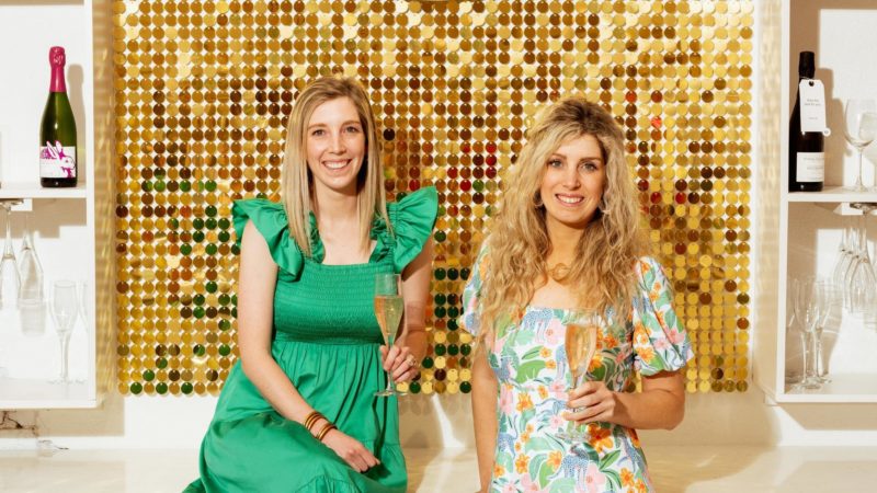 Danielle and Mary Prestifilippo sitting on a ledge in front of the sign inside their boutique